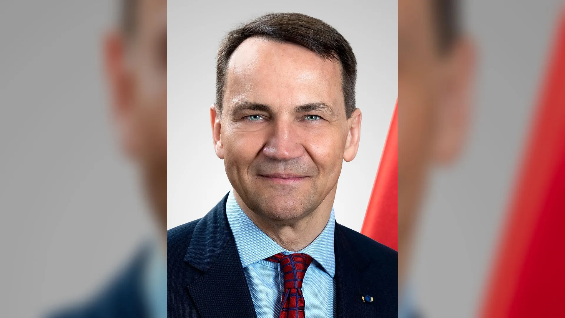Lacivert ceketli, kırmızı kravatlı gülümseyen kısa saçlı adam, arkada kırmızı bayrak