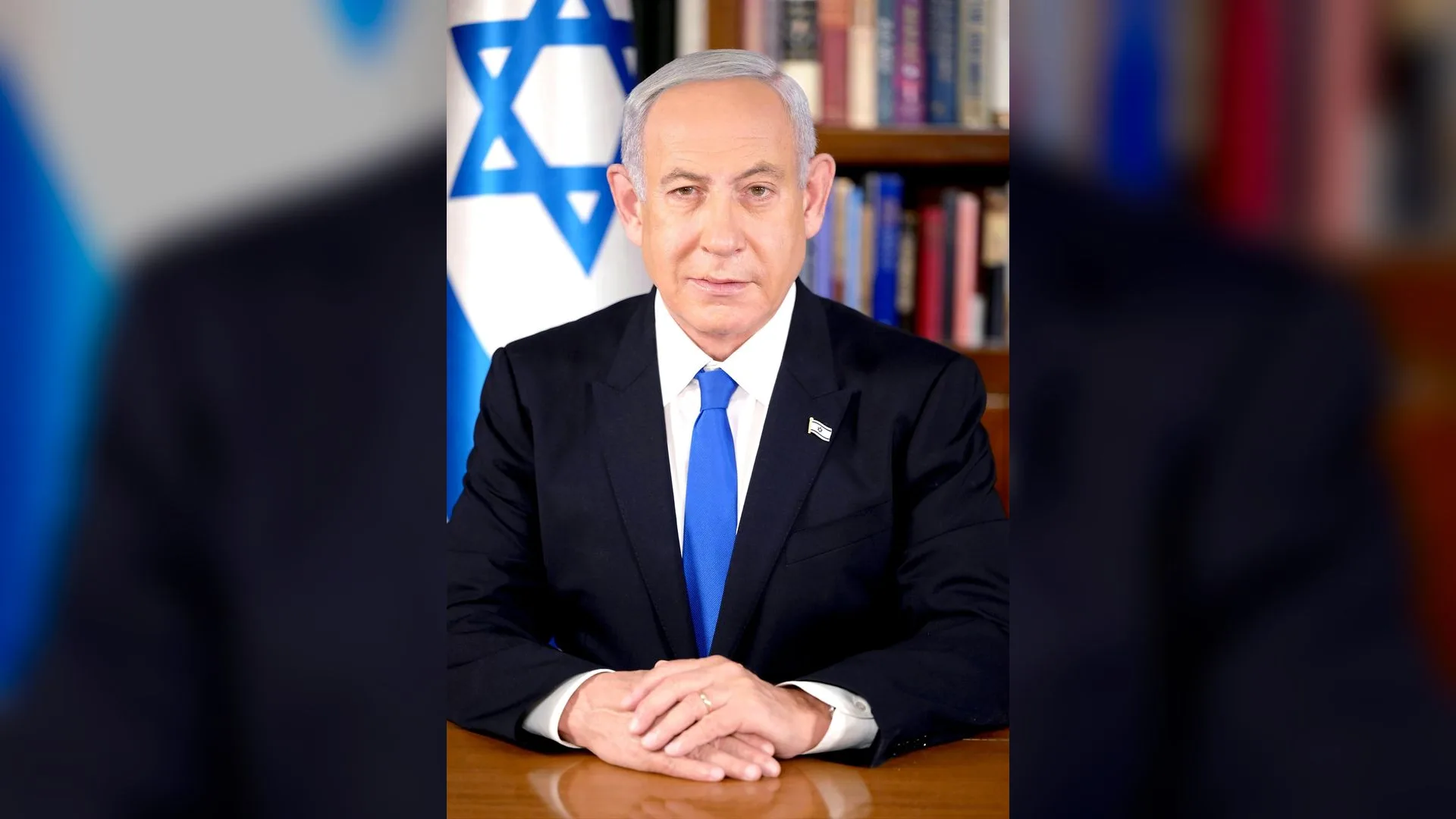 Mavi kravatlı, gri saçlı bir adam İsrail bayrağı ve kitaplık önünde masada oturuyor