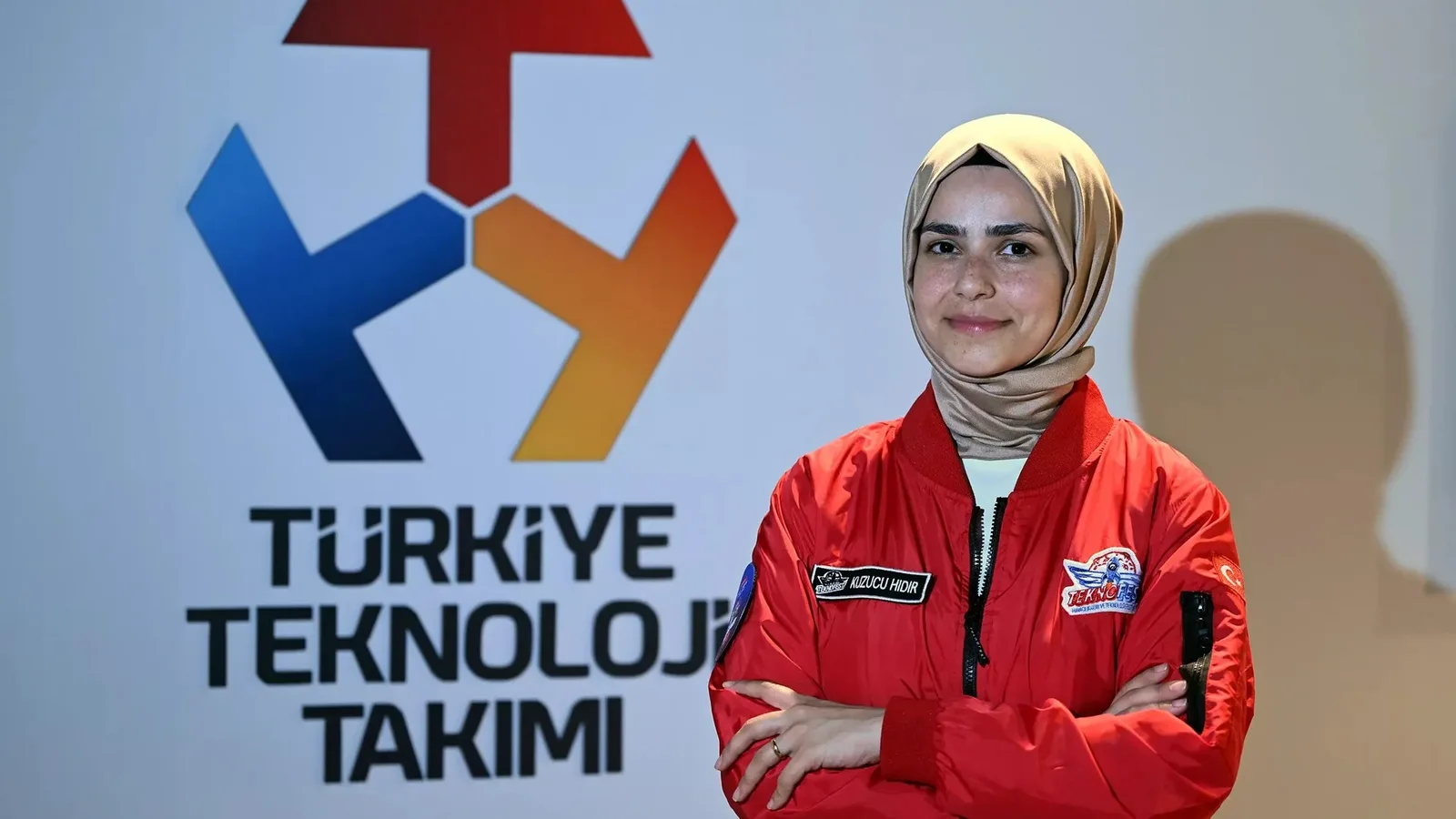 Türkiye Teknoloji Takımı logosu yanında kırmızı ceketi takımlı başörtülü kadın.