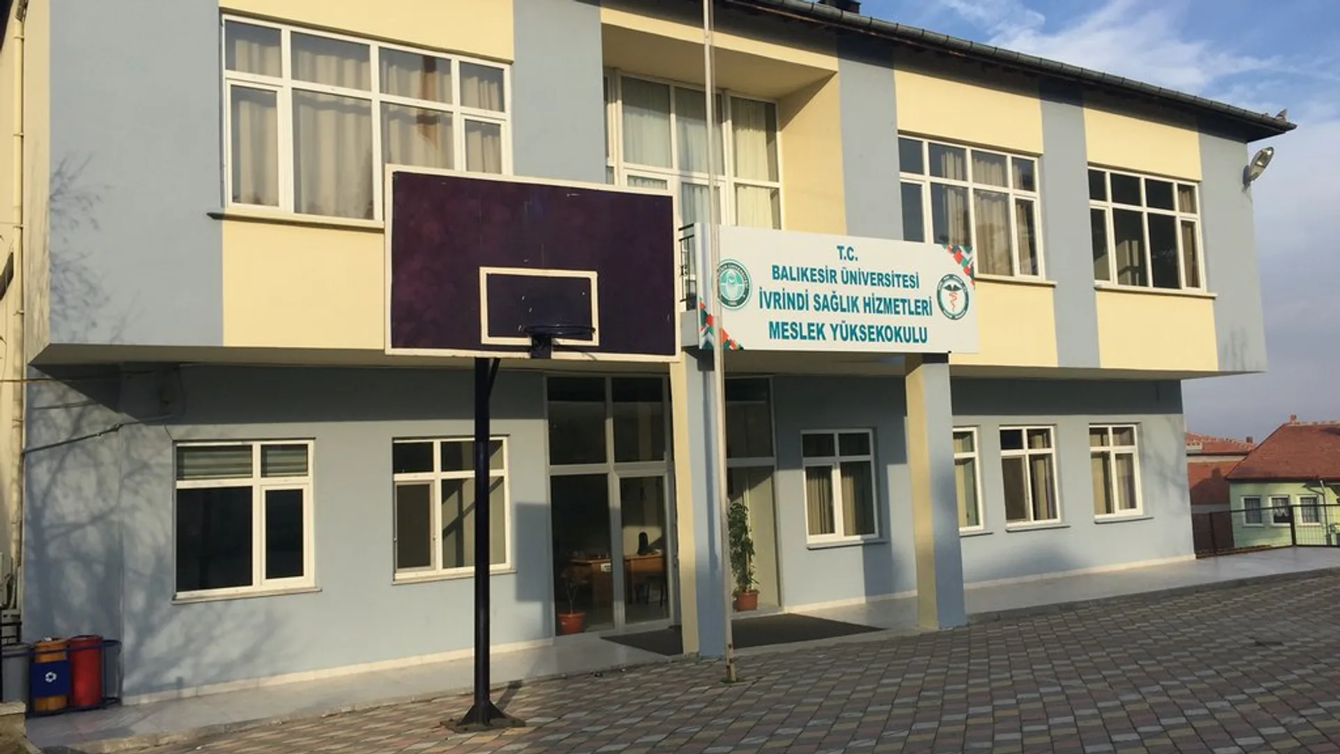 Tabelasında İvrindi Sağlık Hizmetleri MYO yazan iki katlı bina, önünde basketbol potası ve bayrak direği