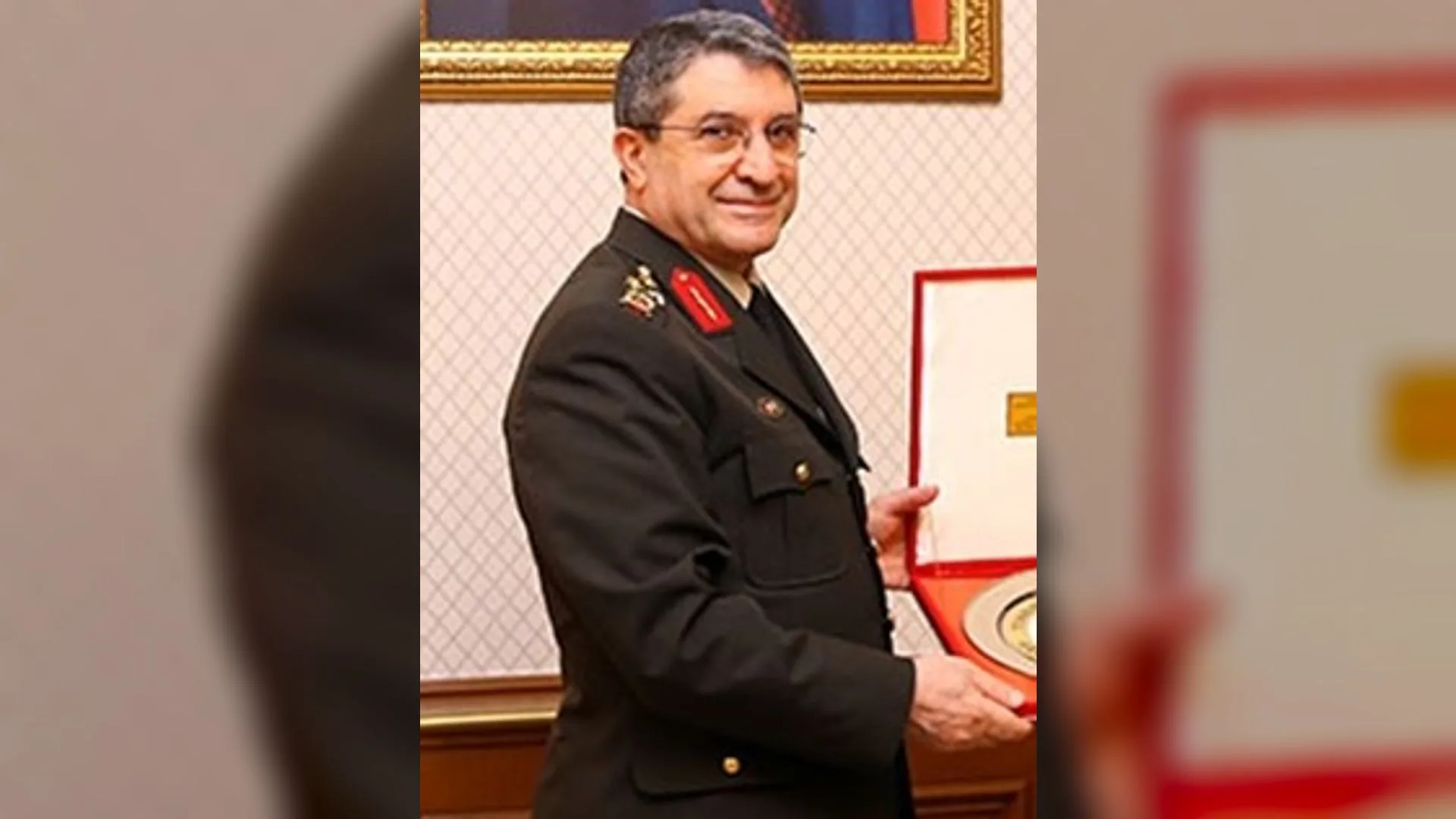 Gözlüklü, gülümseyen bir general elinde kırmızı kutulu bir plaketle duruyor