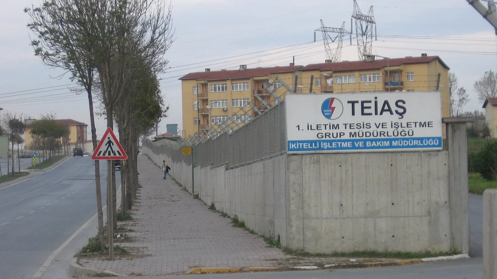 Betonlu duvar üzerinde TEİAŞ şirket logosu ve tabelası, arkada sarı-turuncu bina.