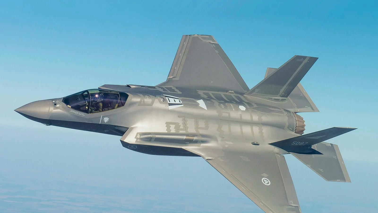 F-35 savaş uçağı gökyüzünde yan görünümde, açık mavi arka plan.