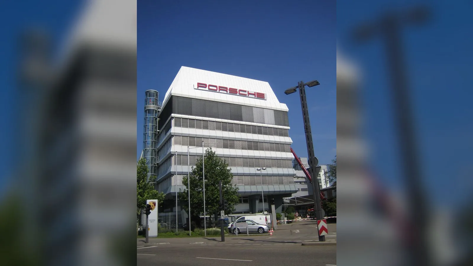 Porsche logolu çok katlı beyaz binası, yeşil alanlı modern şehirsel ortamda.