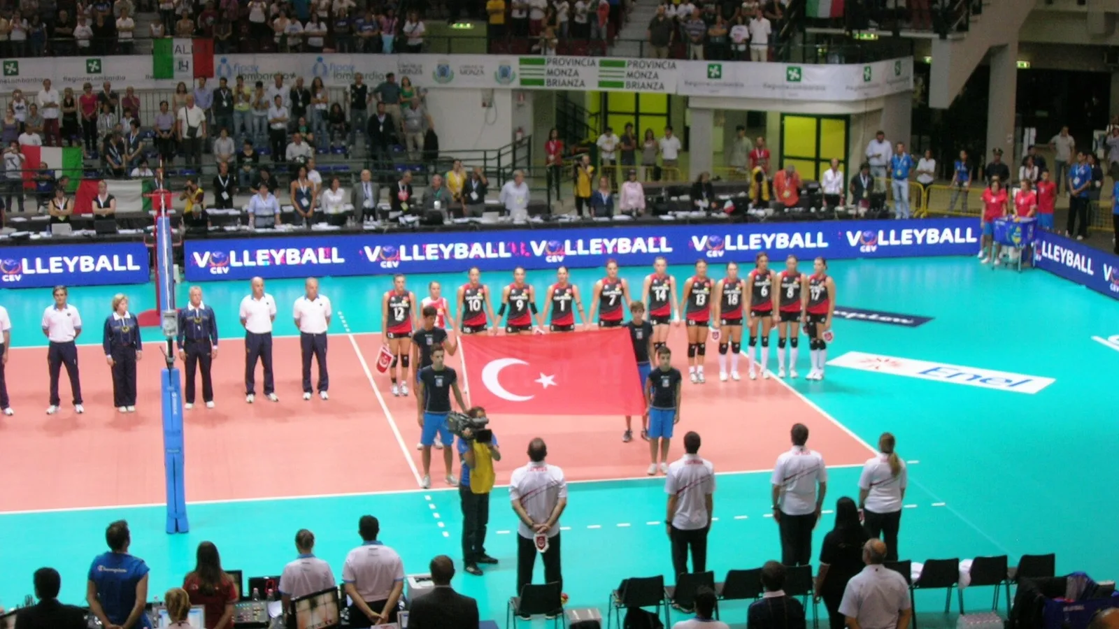 Voleybol sahası, Türk bayrağı, kırmızı formaylı kadın oyuncular ve masanın başında maç yöneticileri.