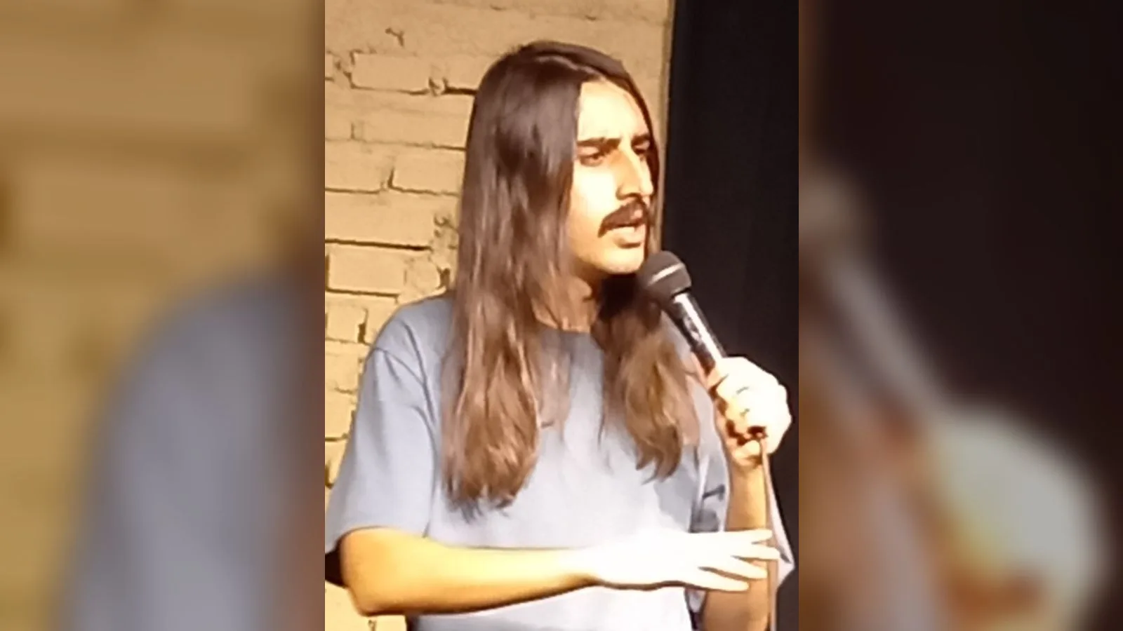 Mikrofon tutan, uzun koyu saçlı erkek, tuğla duvar önünde.