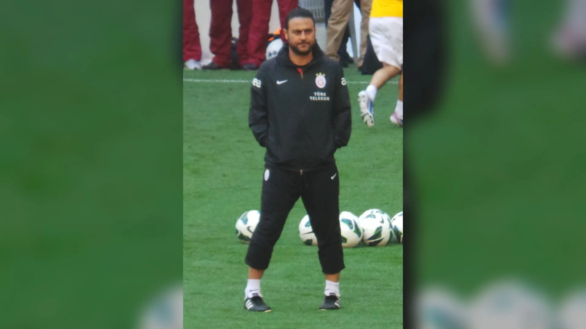 Siyah eşofmanlı sakallı bir adam çimde duruyor, arkasında futbol topları var