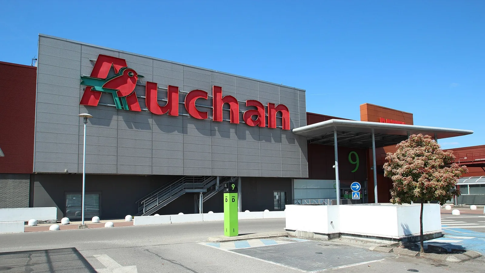 Kırmızı Auchan logosu bulunan gri cephe, alışveriş merkezi binası, açık havada.