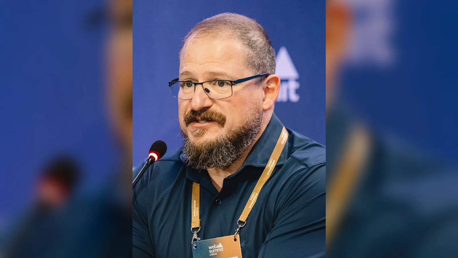 Gözlüklü, bıyıklı erkek konferans masasında mikrofon başında.