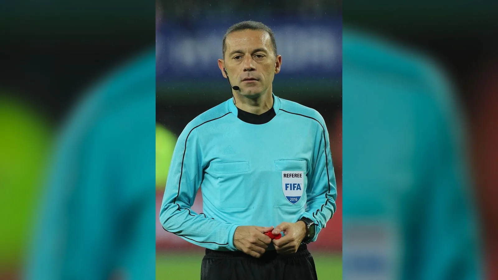 Açık mavi hakem forması giymiş orta yaşlı kişi futbol sahasında.