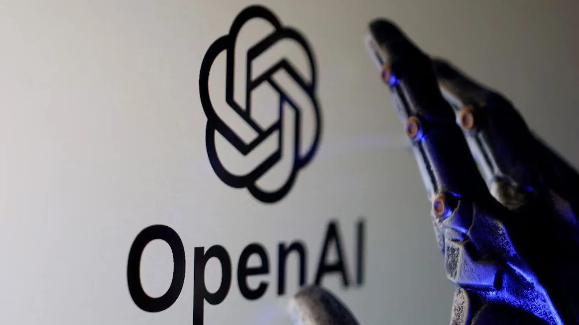 Beyaz duvarda siyah OpenAI logosu ve yazısı, önünde mavi ışıkla aydınlanan robot eli