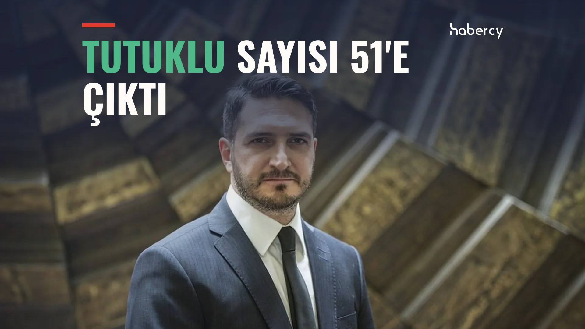 Takım elbiseli, kravatlı, sakallı bir adam taş duvar önünde poz veriyor