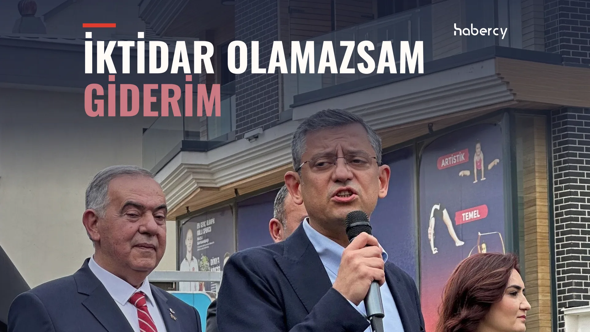 Takım elbiseli gözlüklü bir adam mikrofonla konuşuyor, yanında kırmızı kravatlı yaşlı bir adam duruyor
