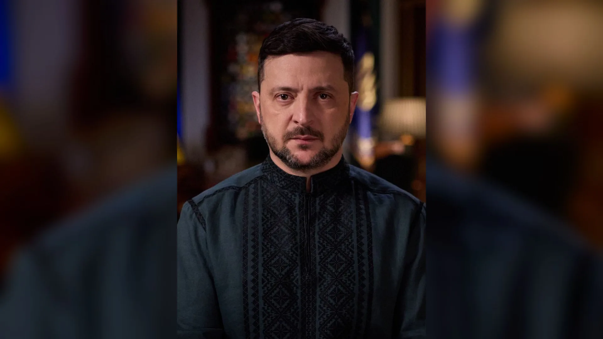 İşlemeli koyu yeşil gömlekli, sakallı bir adam kameraya bakıyor