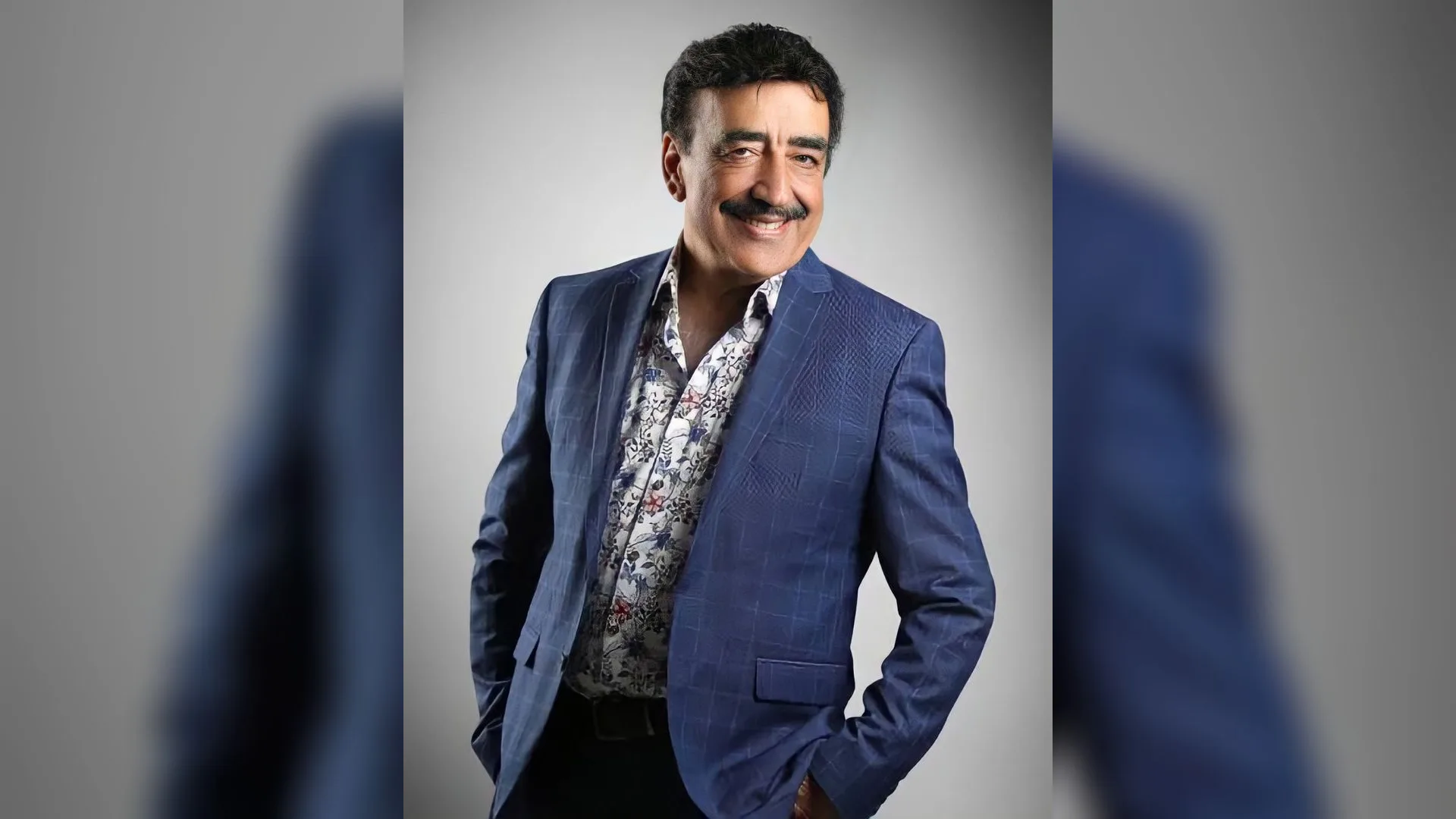 Mavi ceketli, desenli gömlekli, bıyıklı ve gülümseyen bir adam gri fon önünde poz veriyor