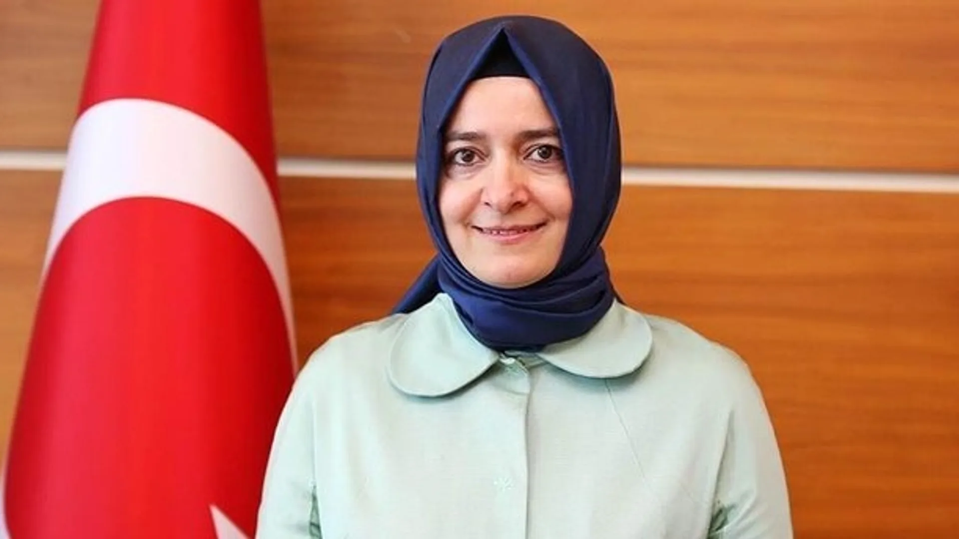 Türk bayrağı önünde lacivert başörtülü, açık yeşil ceketli gülümseyen bir kadın
