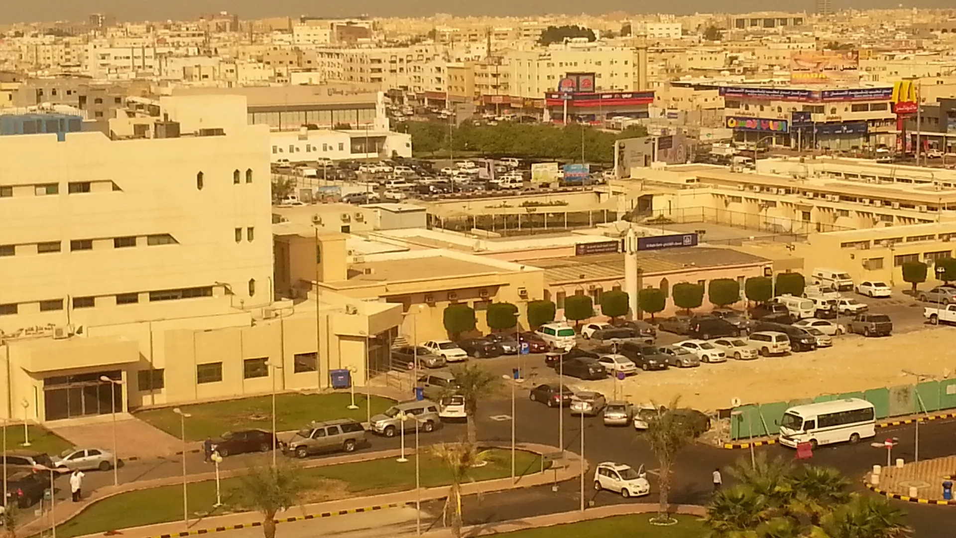 Tozlu havada bej binalar, otoparklar ve palmiyelerle çevrili bir şehir manzarası