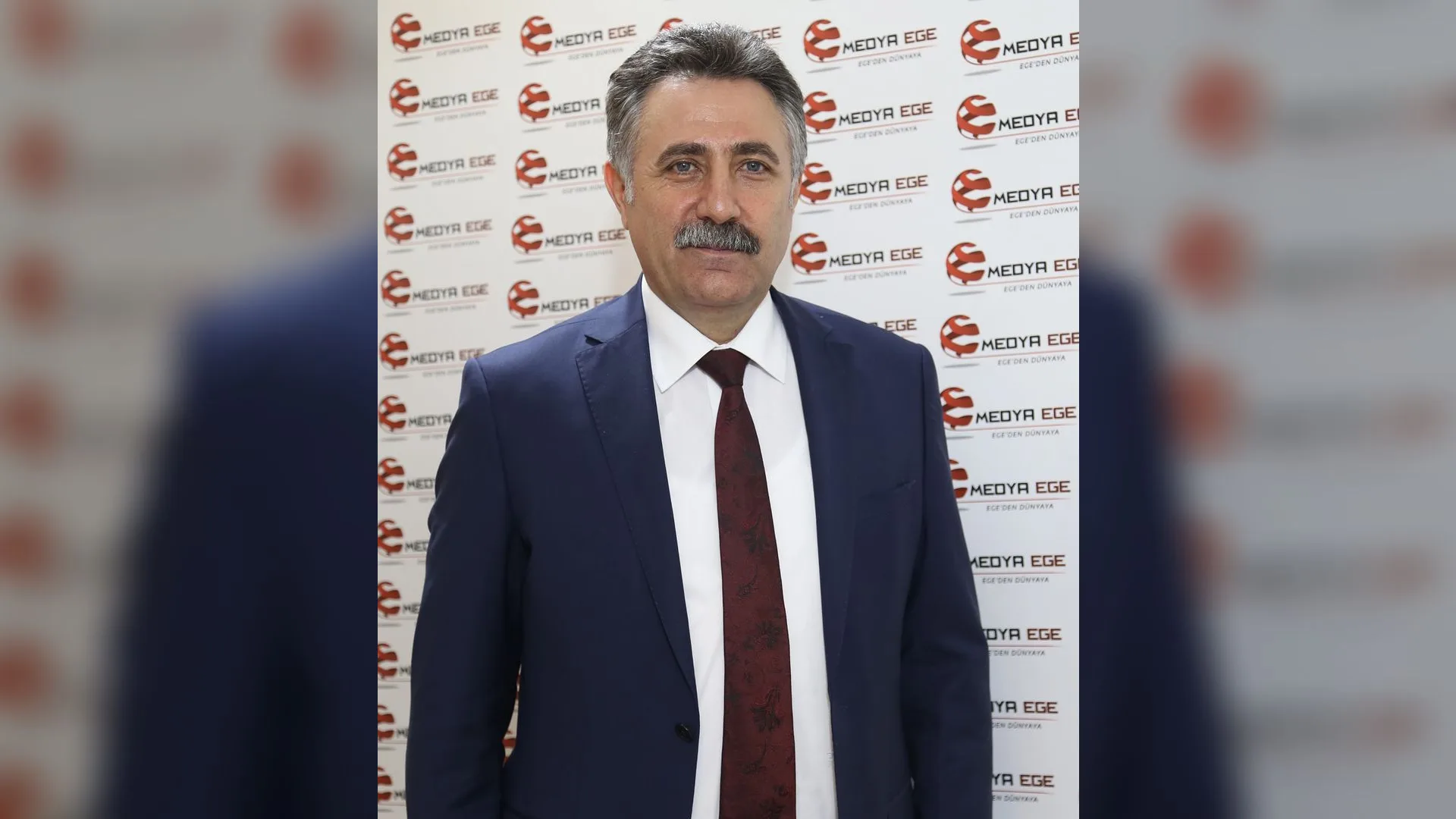 Logolu panonun önünde lacivert takım elbiseli, bordo kravatlı, bıyıklı gri saçlı bir adam