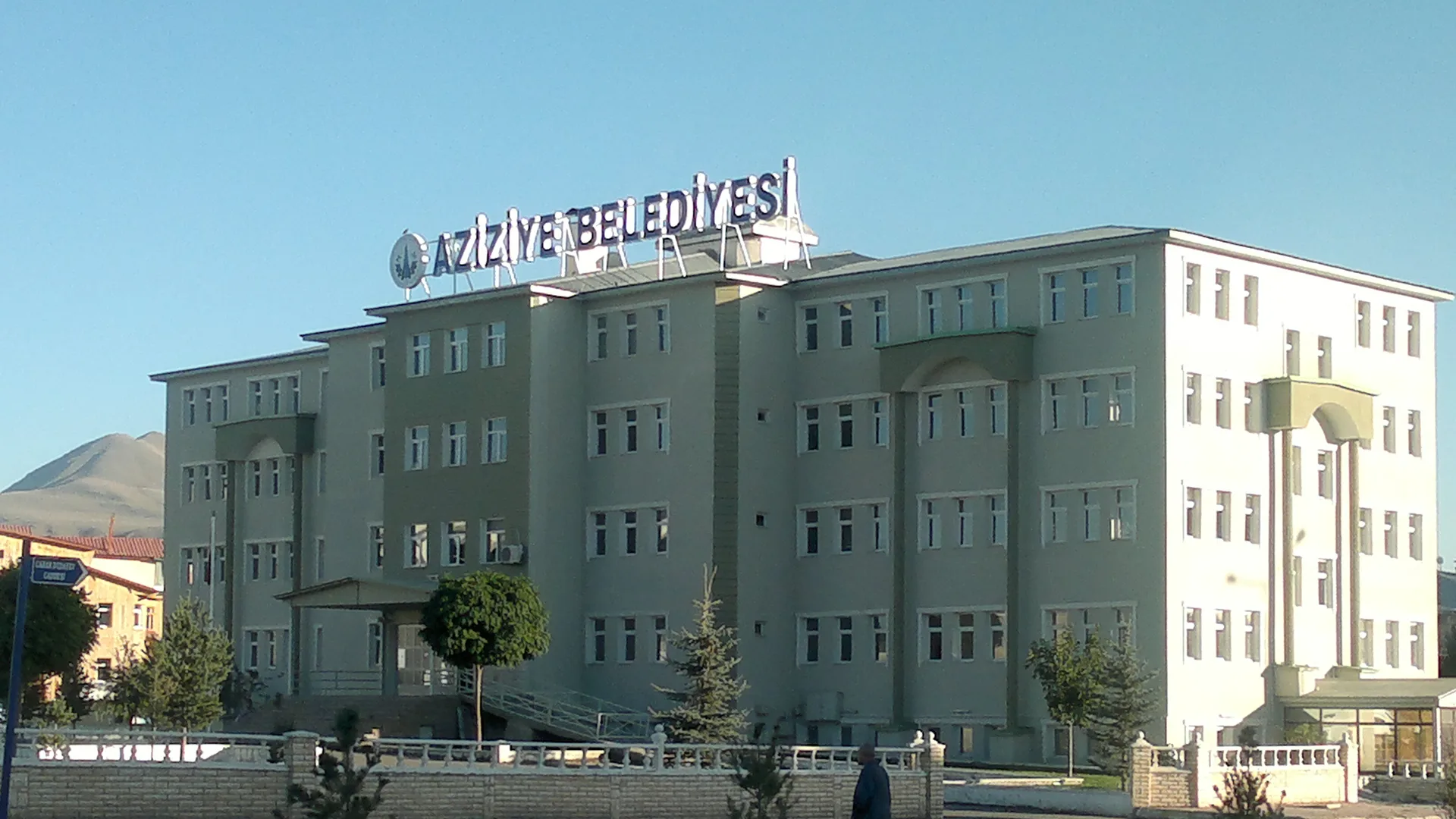 Çatısında Aziziye Belediyesi yazan çok katlı gri bina ve önündeki yolda yürüyen bir kişi
