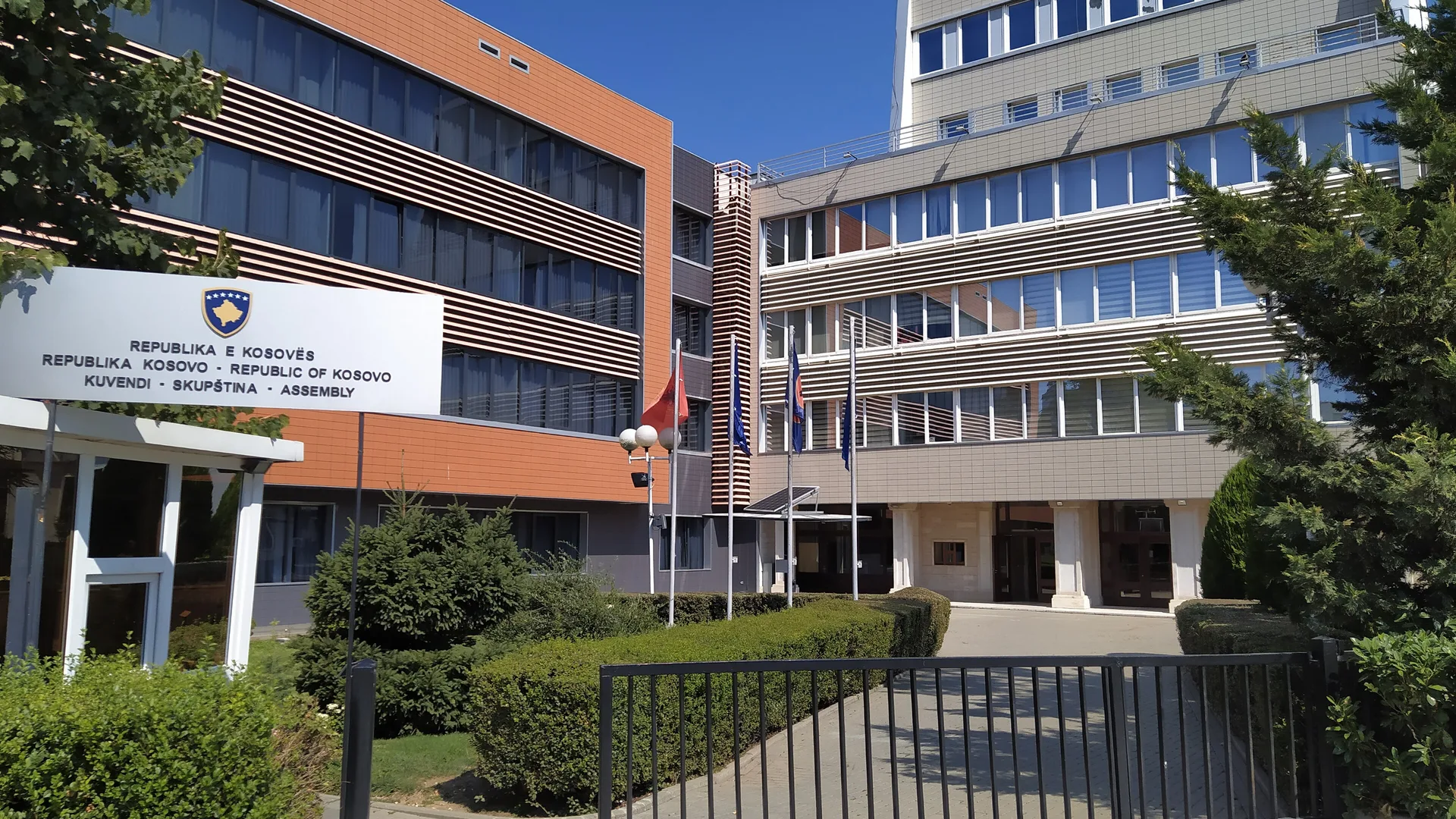 Önünde Kosova Meclisi tabelası ve bayraklar bulunan modern bir binanın girişi