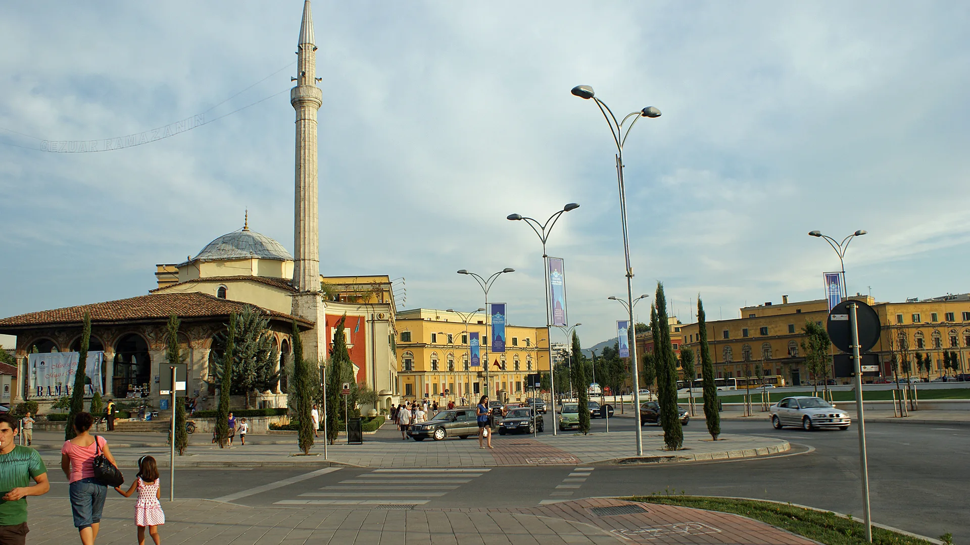 Minareli kubbeli bir cami, sarı binalar ve yayaların yürüdüğü geniş bir meydan