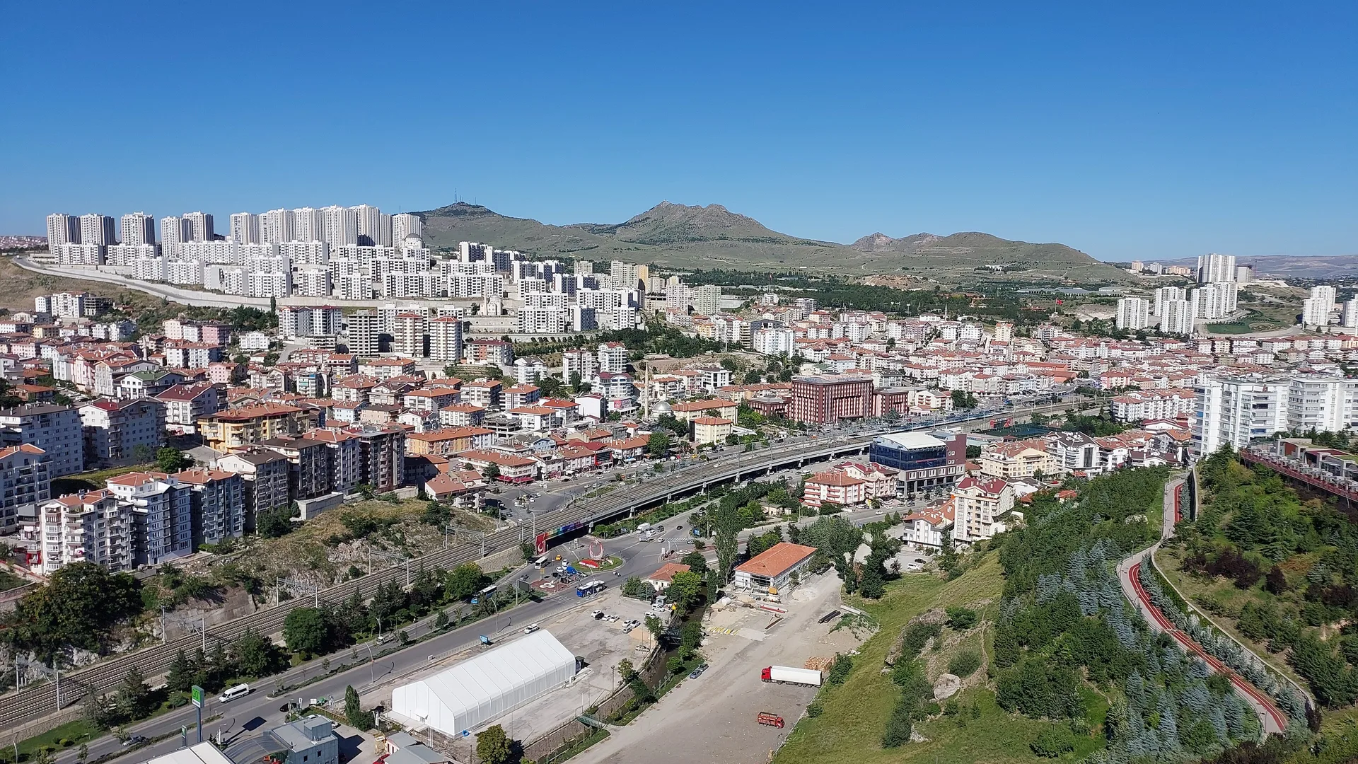 Yüksekten çekilmiş, apartmanlar, geniş bir yol ve arkada tepelerin göründüğü bir şehir manzarası