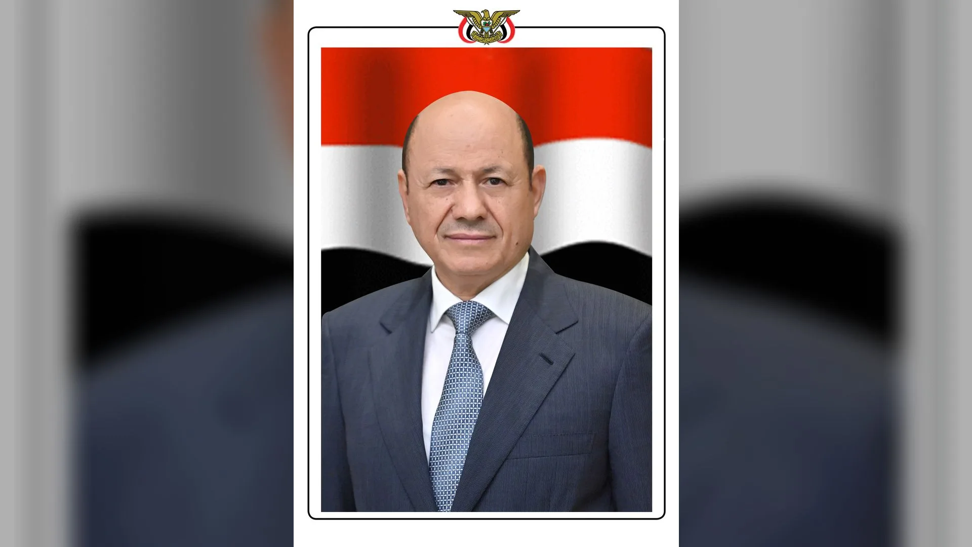 Yemen bayrağı önünde lacivert takım elbiseli, kravatlı kel bir adamın resmi portresi