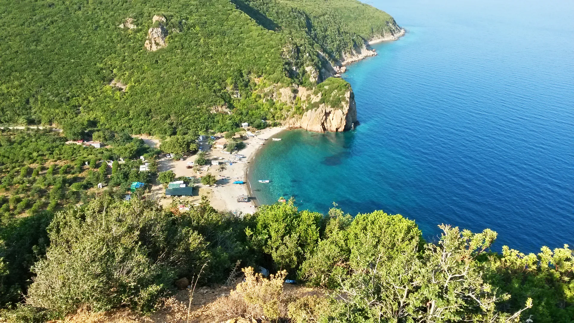 Yeşil ormanlık tepelerin arasında turkuaz denize açılan kumsallı bir koy ve kıyıdaki küçük tekneler