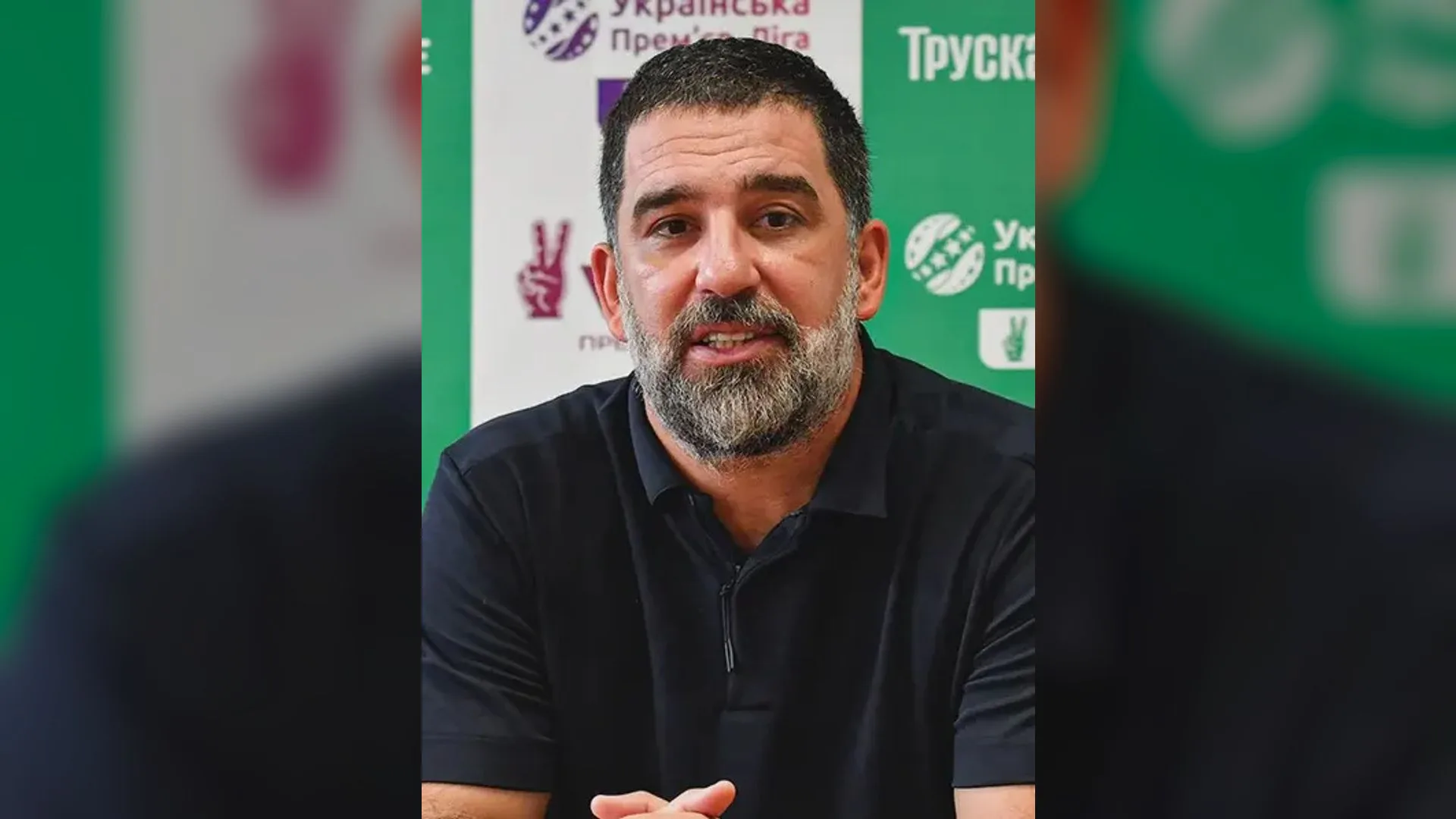 Sakallı, siyah tişörtlü bir adam yeşil sponsor panosu önünde konuşuyor