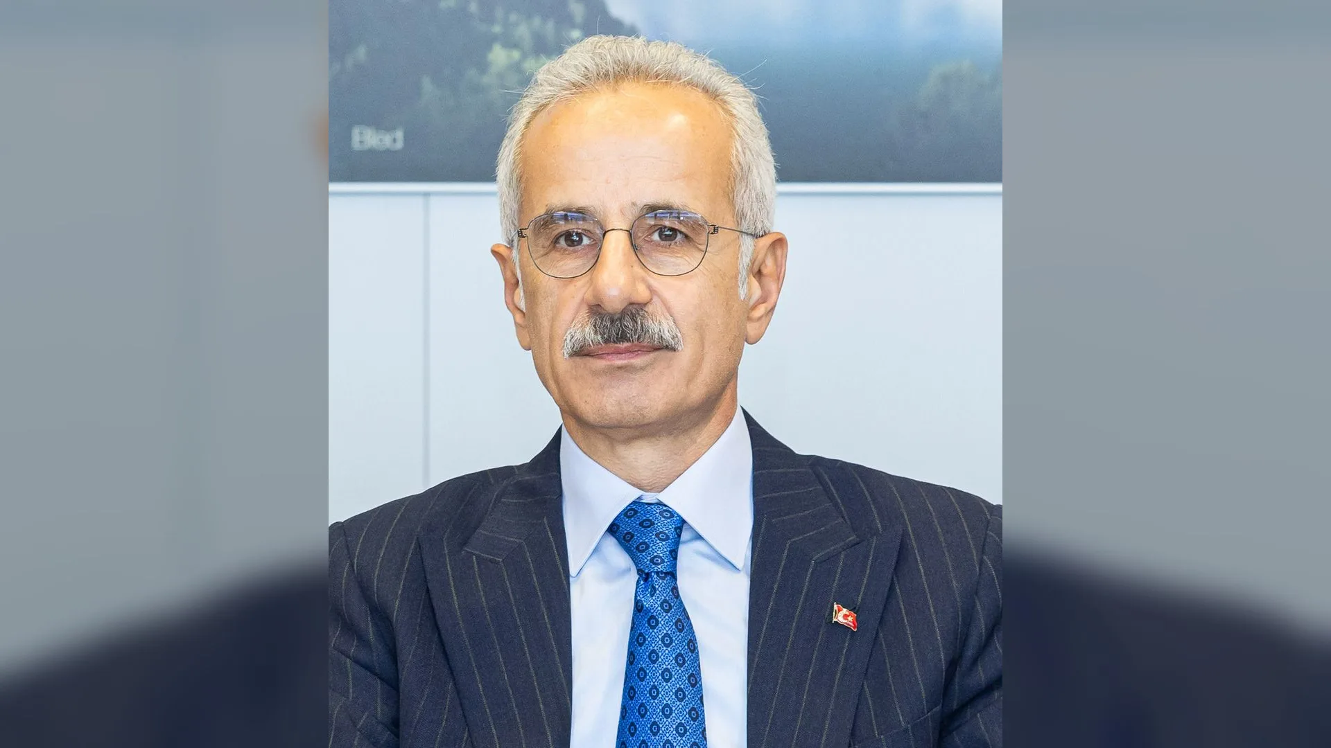 Gözlüklü, bıyıklı, beyaz saçlı, lacivert çizgili takım elbise ve mavi kravat giyen bir adam