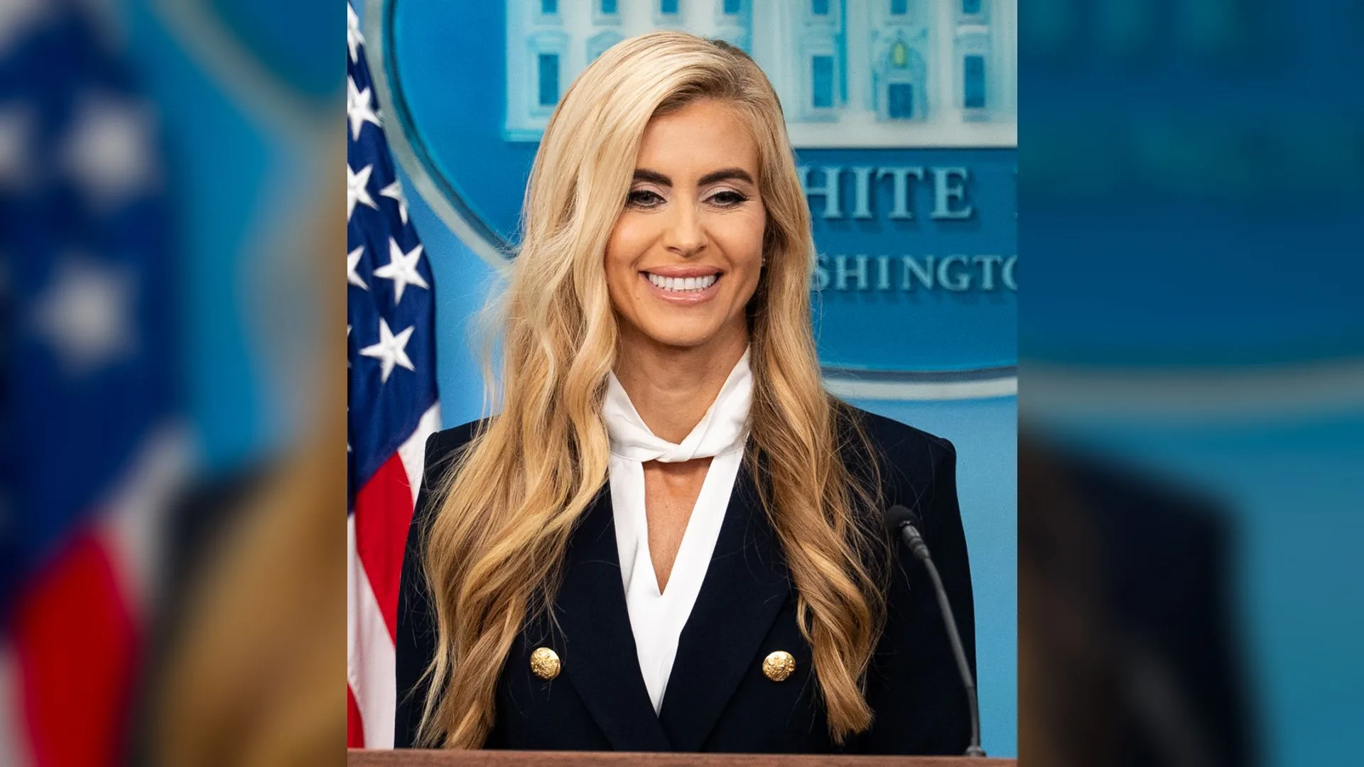 Uzun sarı saçlı, lacivert ceketli bir kadın, Amerikan bayrağı önünde kürsüde gülümsüyor.