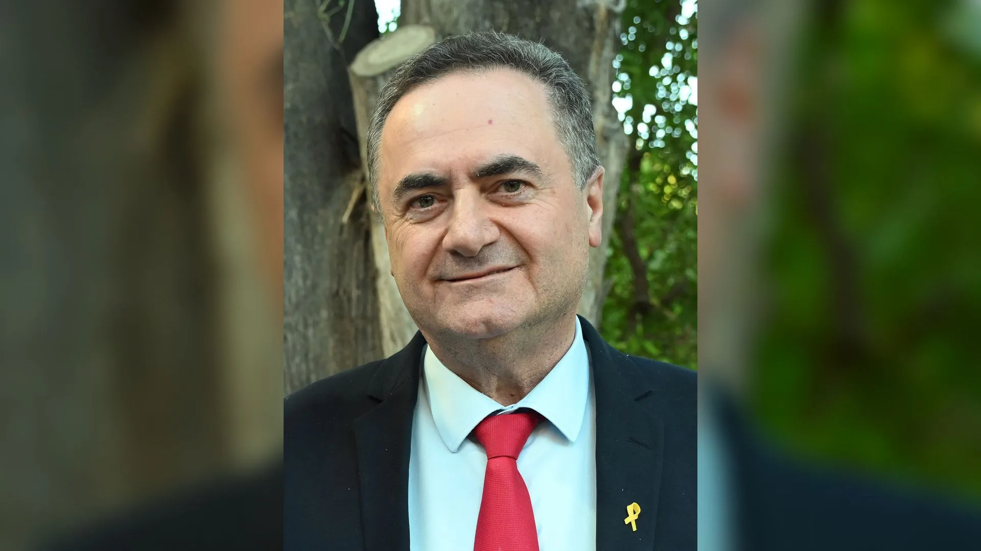 Lacivert ceketli, kırmızı kravatlı, yakasında sarı kurdele olan gülümseyen adam, arkada ağaçlar