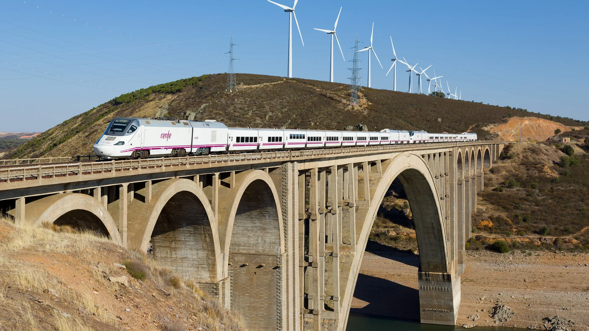 Beyaz-mor Renfe hızlı treni, arkasında rüzgâr türbinleri olan taş kemerli viyadükten geçiyor