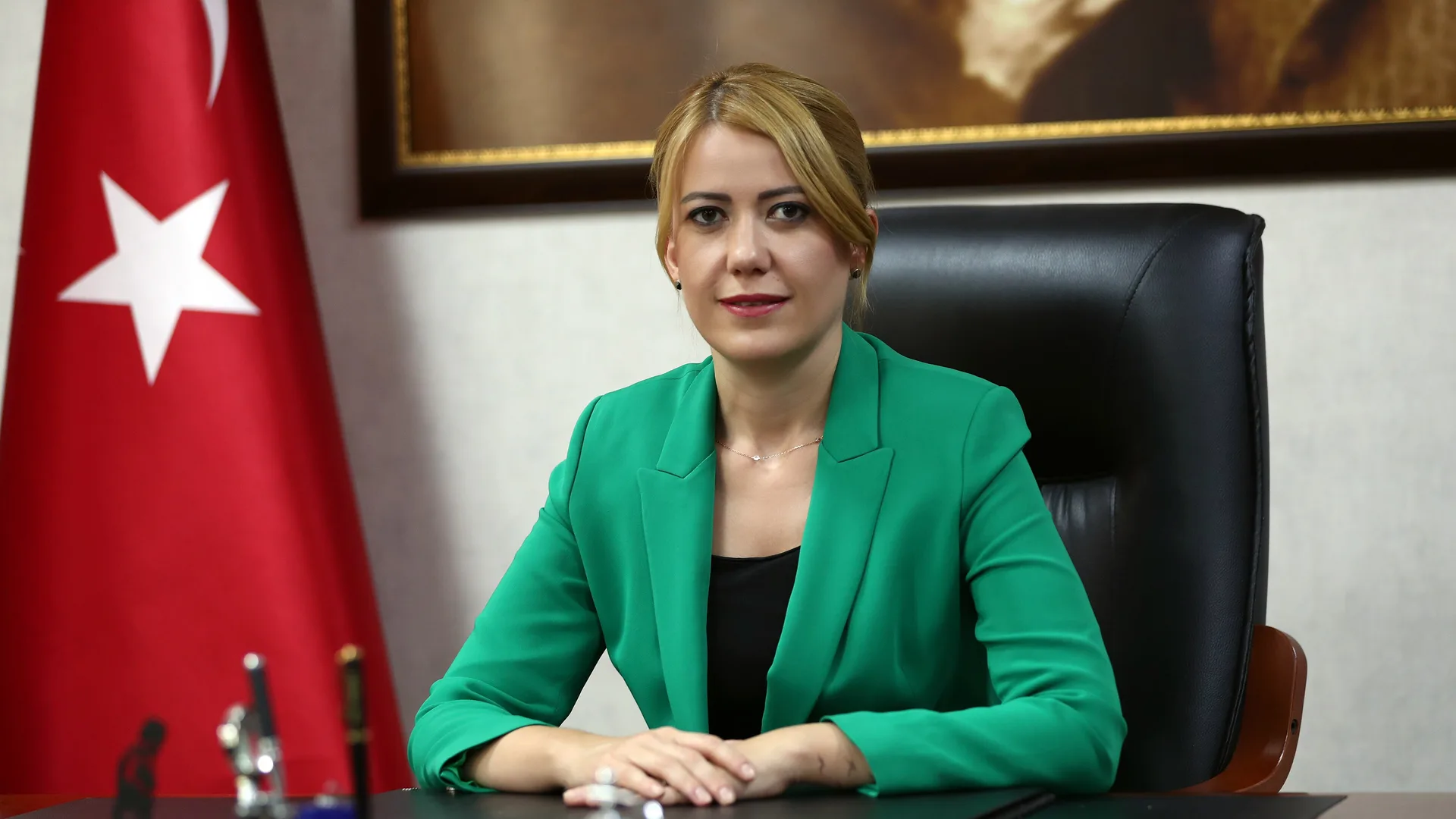 Türk bayrağı önünde masada oturan yeşil ceketli sarışın bir kadın