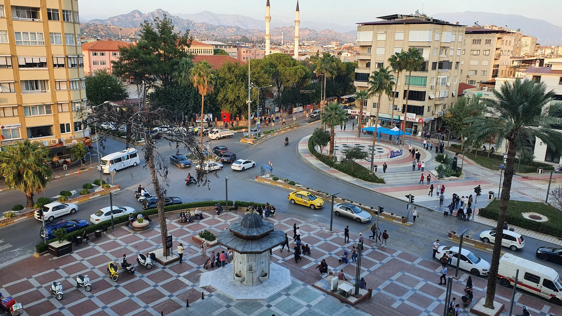 Palmiyeli bir kent meydanı, ortada kubbeli çeşme, arkada çift minareli cami ve dağlar