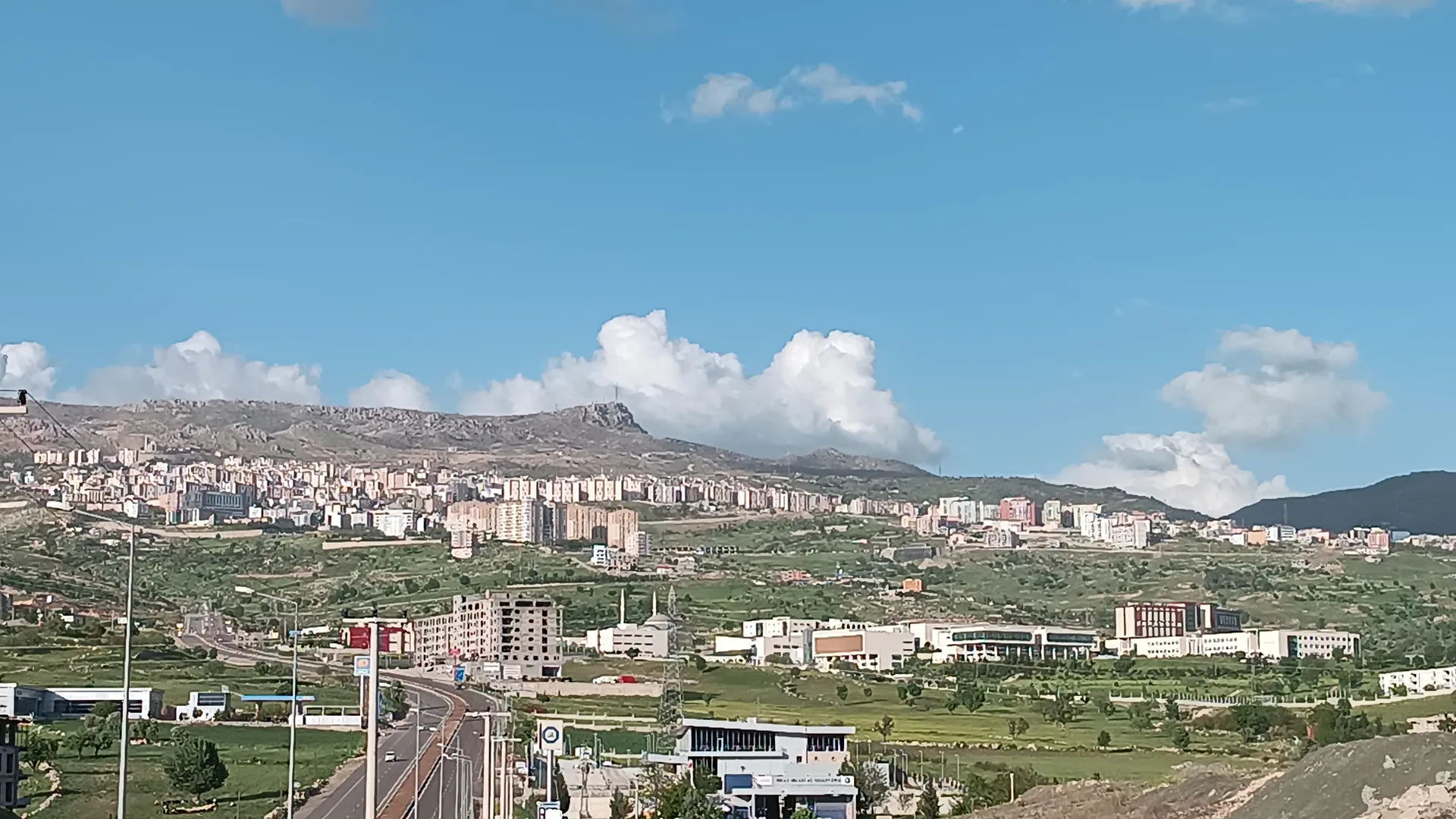 Dağ eteğine yayılmış apartmanlar, yeşil düzlük ve geniş bir yol, mavi gökyüzü
