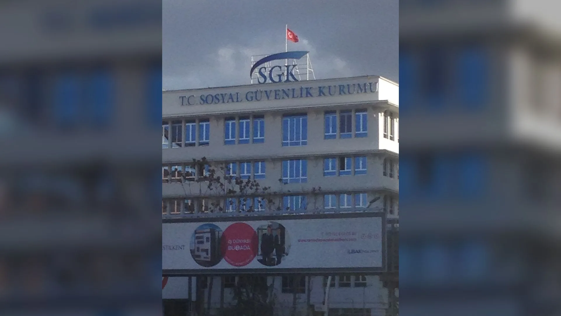 Çatısında SGK logosu ve Türk bayrağı bulunan Sosyal Güvenlik Kurumu binası, önünde reklam panosu