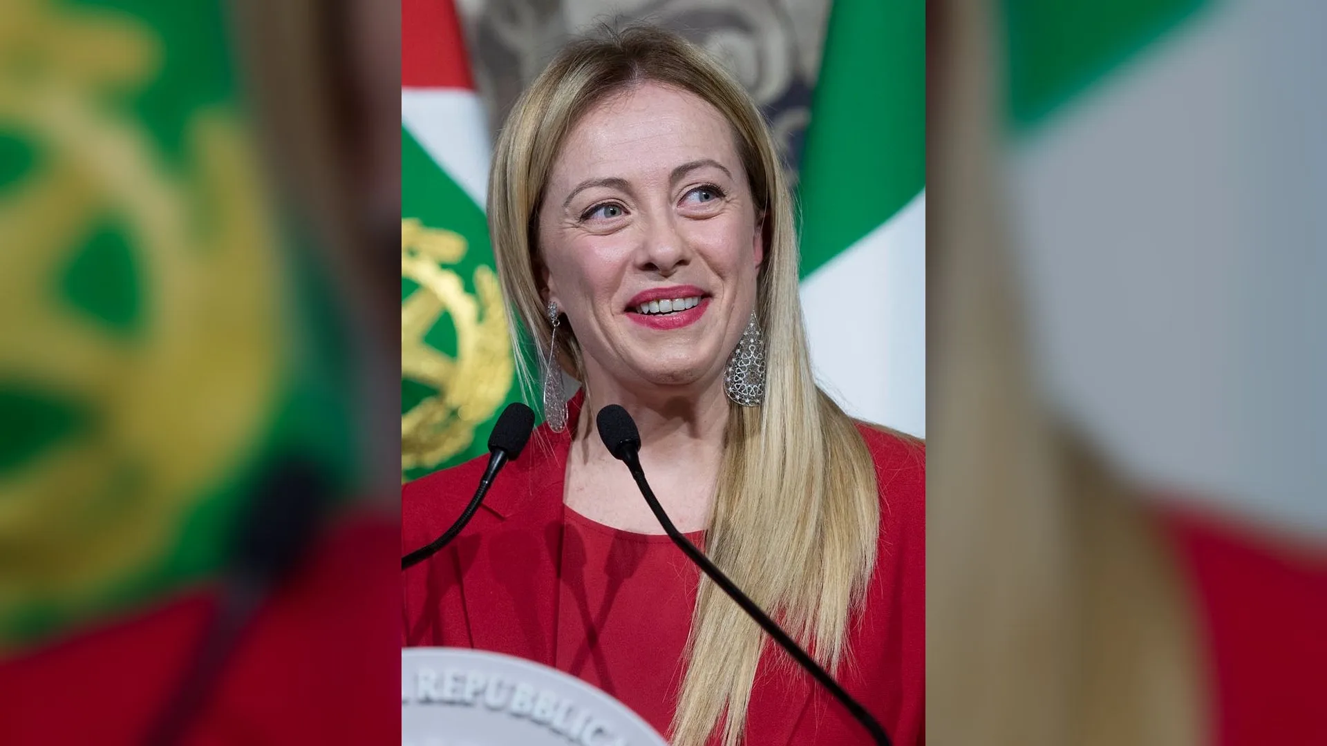 Kırmızı giysili sarışın bir kadın, İtalyan bayrağı önünde mikrofonlu kürsüde gülümsüyor.