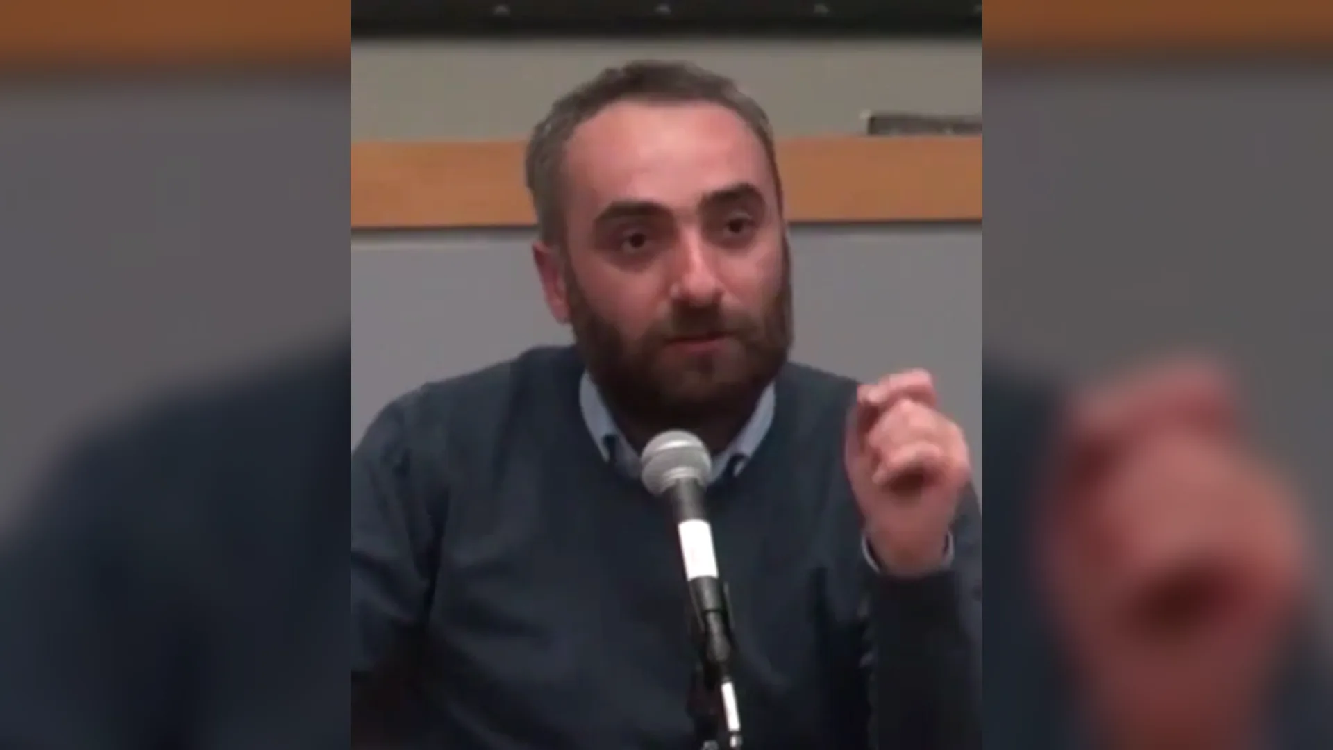Sakallı, lacivert kazaklı bir adam mikrofona konuşurken elini kaldırıyor