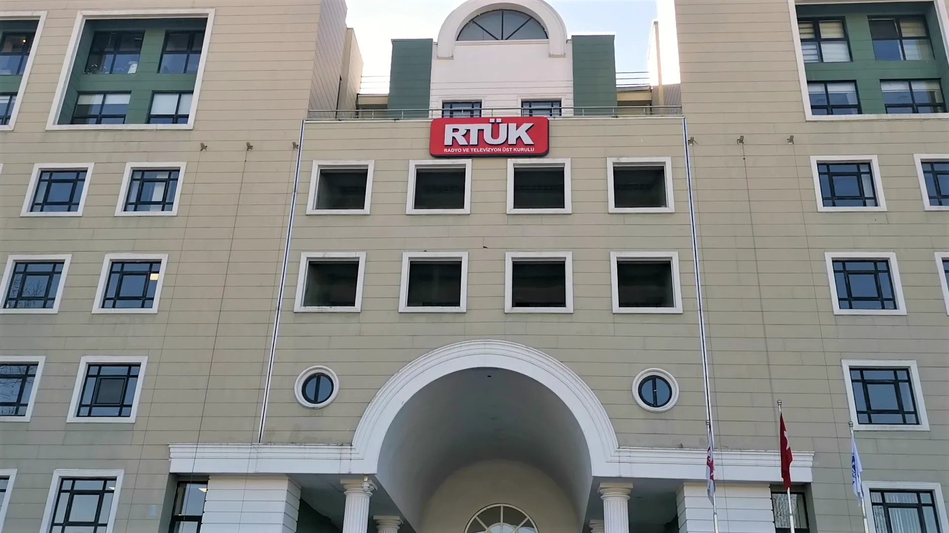 Bej renkli çok katlı bir binanın cephesinde kırmızı RTÜK tabelası, kemerli giriş ve bayraklar