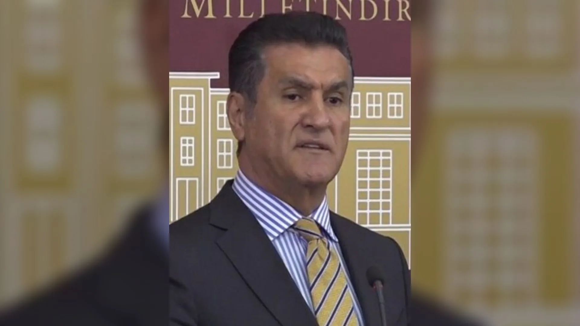 Meclis basın odasında, çizgili kravatlı ve koyu takım elbiseli gri saçlı bir adam konuşuyor