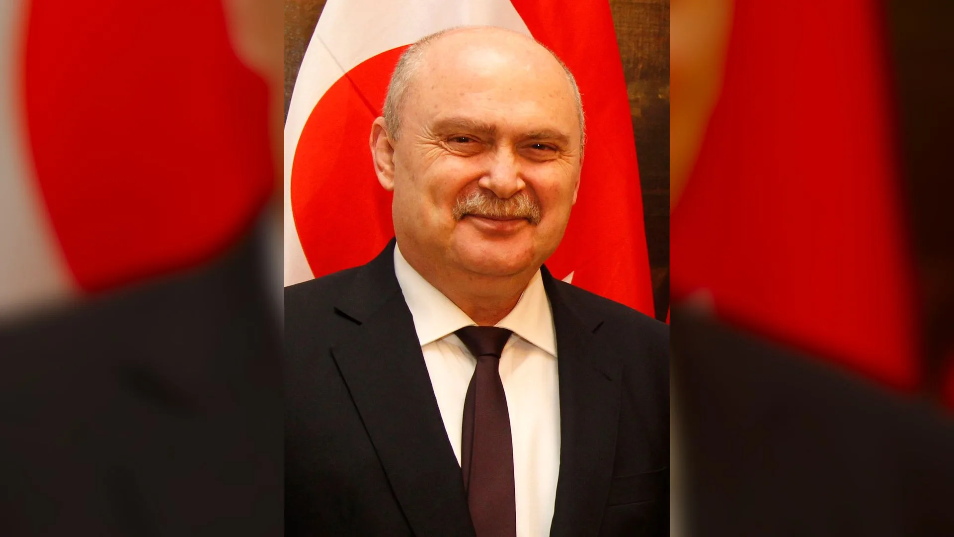 Türk bayrağı önünde gülümseyen, bıyıklı, takım elbiseli kel bir adam