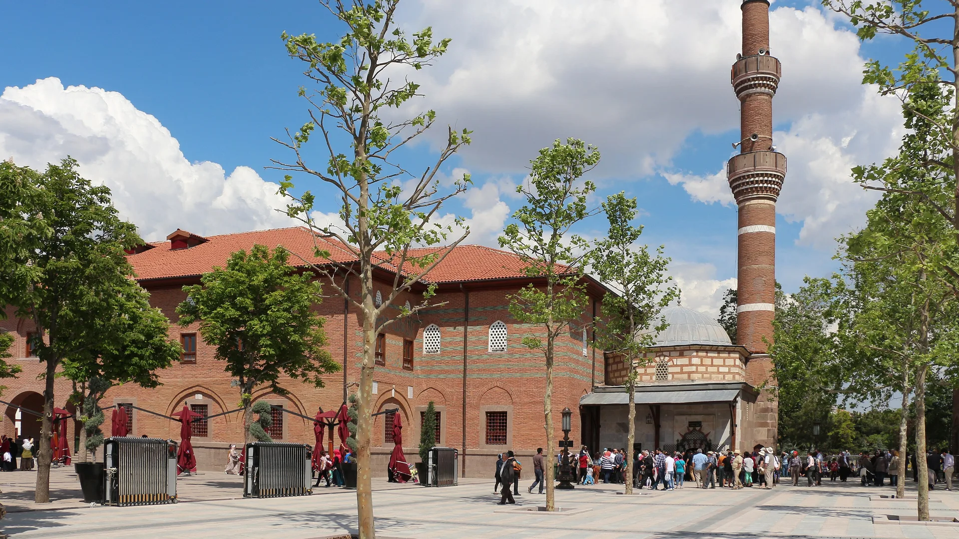 Ağaçlar arasında kırmızı kiremit çatılı tuğla cami ve yüksek tuğla minare, mavi bulutlu gökyüzü