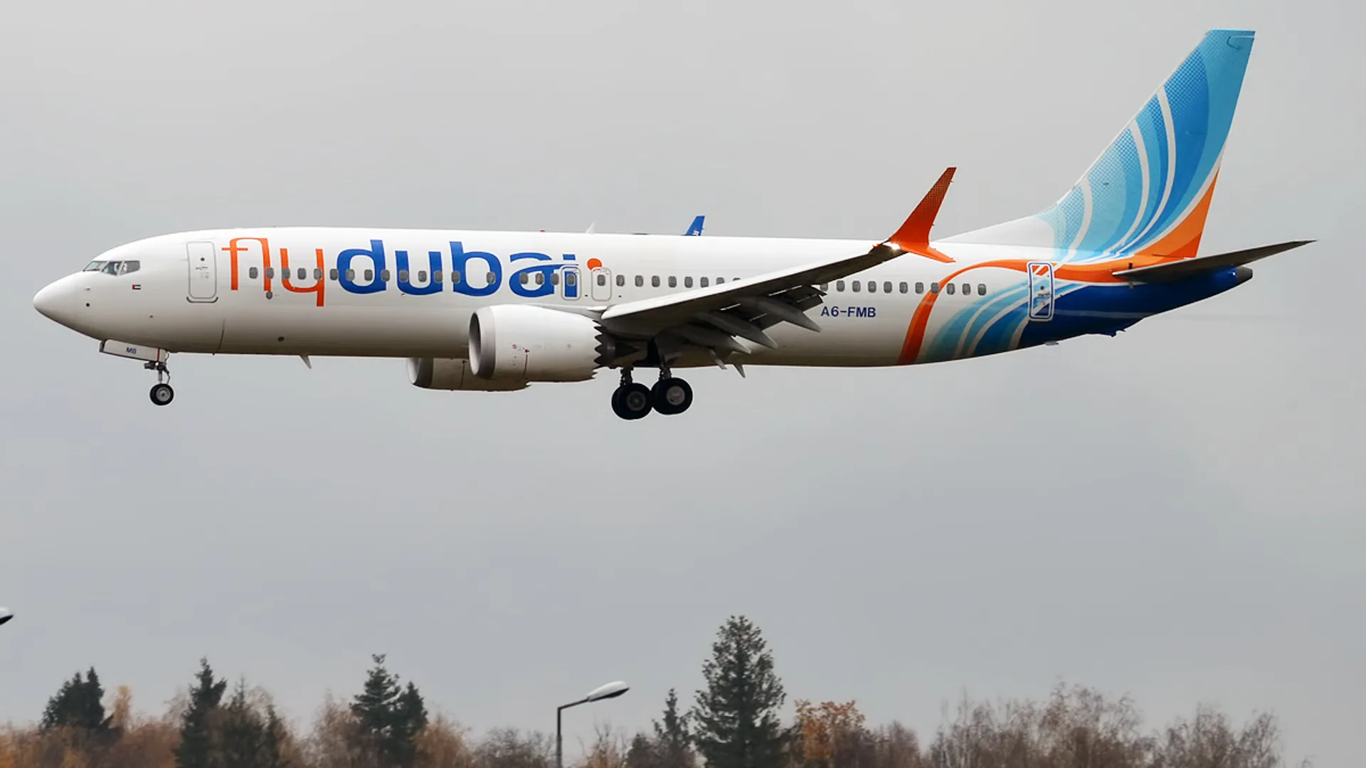 Flydubai logolu beyaz-mavi-turuncu bir yolcu uçağı, açık iniş takımlarıyla gri gökyüzünde uçuyor