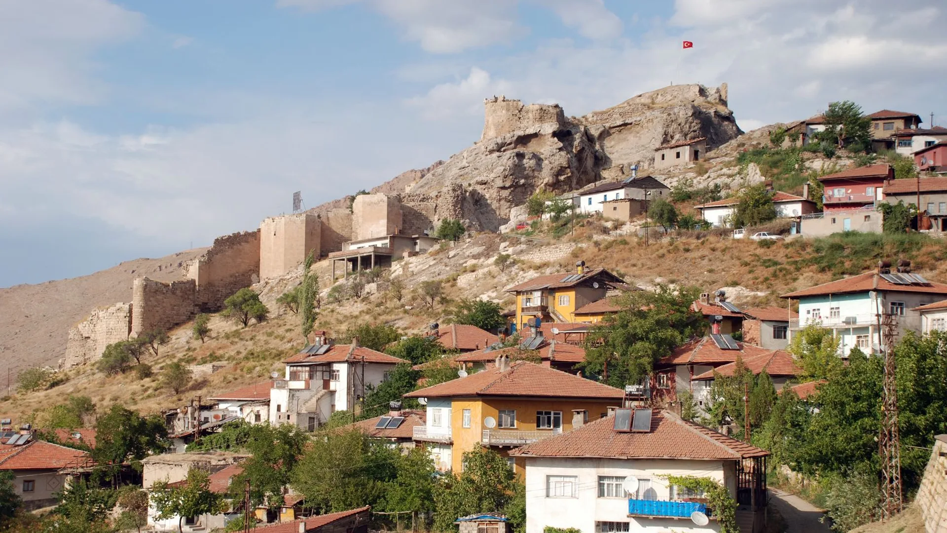 Kayalık tepede kale surları, yamaçta kiremit çatılı renkli evler ve ağaçlar