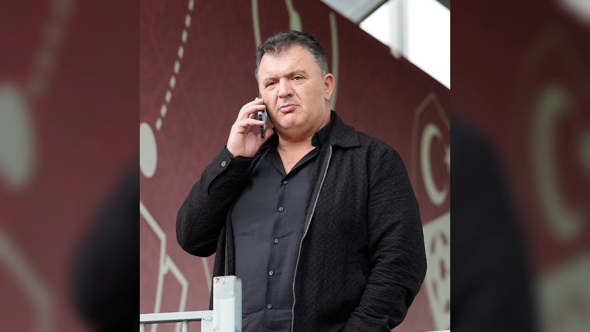 Siyah gömlekli ve ceketli bir adam bordo tribün önünde telefonla konuşuyor