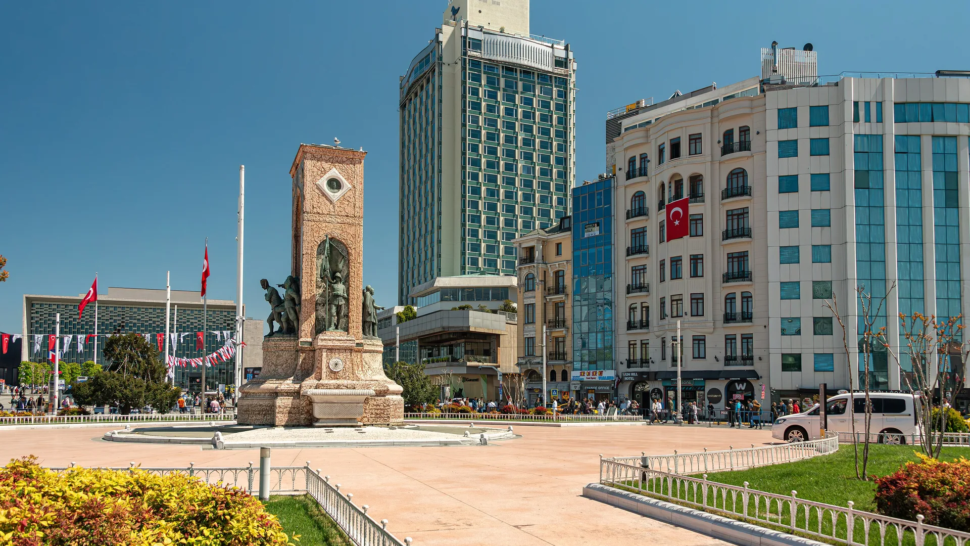 Güneşli bir günde Cumhuriyet Anıtı, bayraklar ve arkada yüksek bir otel kulesiyle geniş bir meydan.