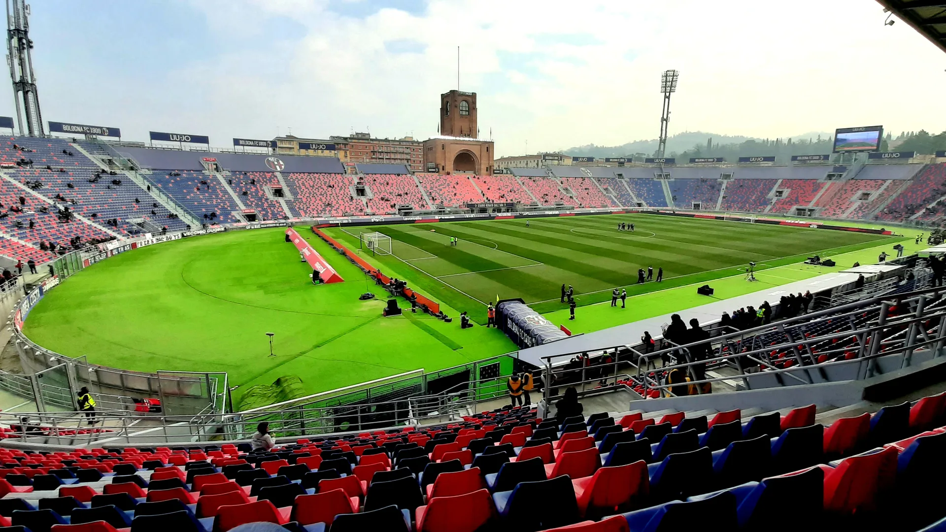 Kırmızı-mavi koltuklu, yeşil sahalı bir futbol stadyumu ve arkada tuğla saat kulesi