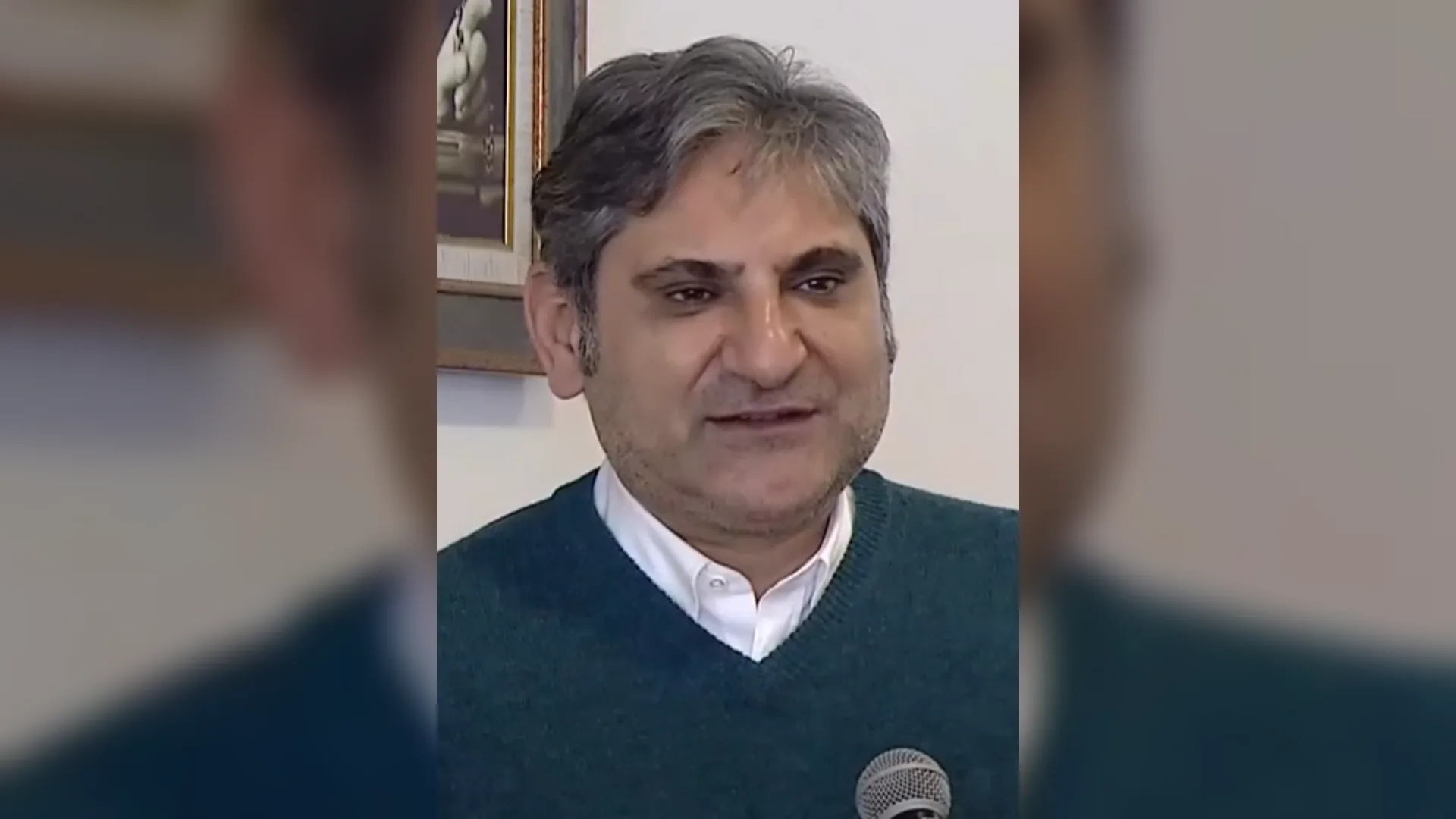 Kır saçlı, yeşil kazak ve beyaz gömlek giymiş bir adam mikrofon önünde gülümseyerek konuşuyor.
