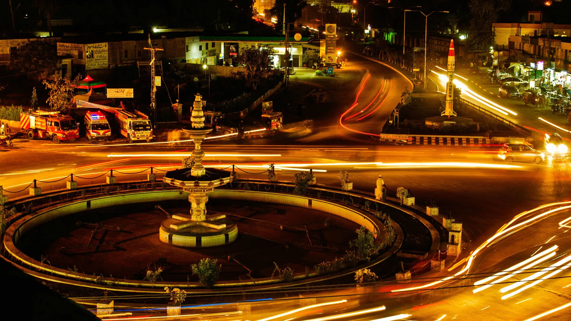 Gece ışıklı bir meydanda çeşme, kenarda park etmiş itfaiye ve ambulanslar, far izleri